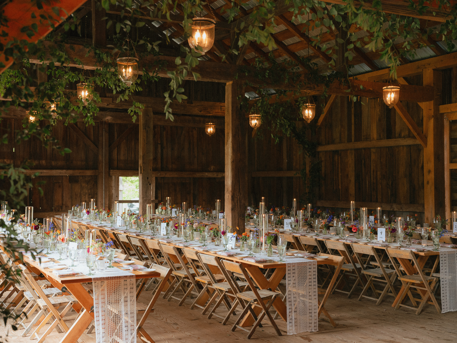 barn wedding reception details with table runners, wooden tables and chairs, and candles, greenery lines the roof