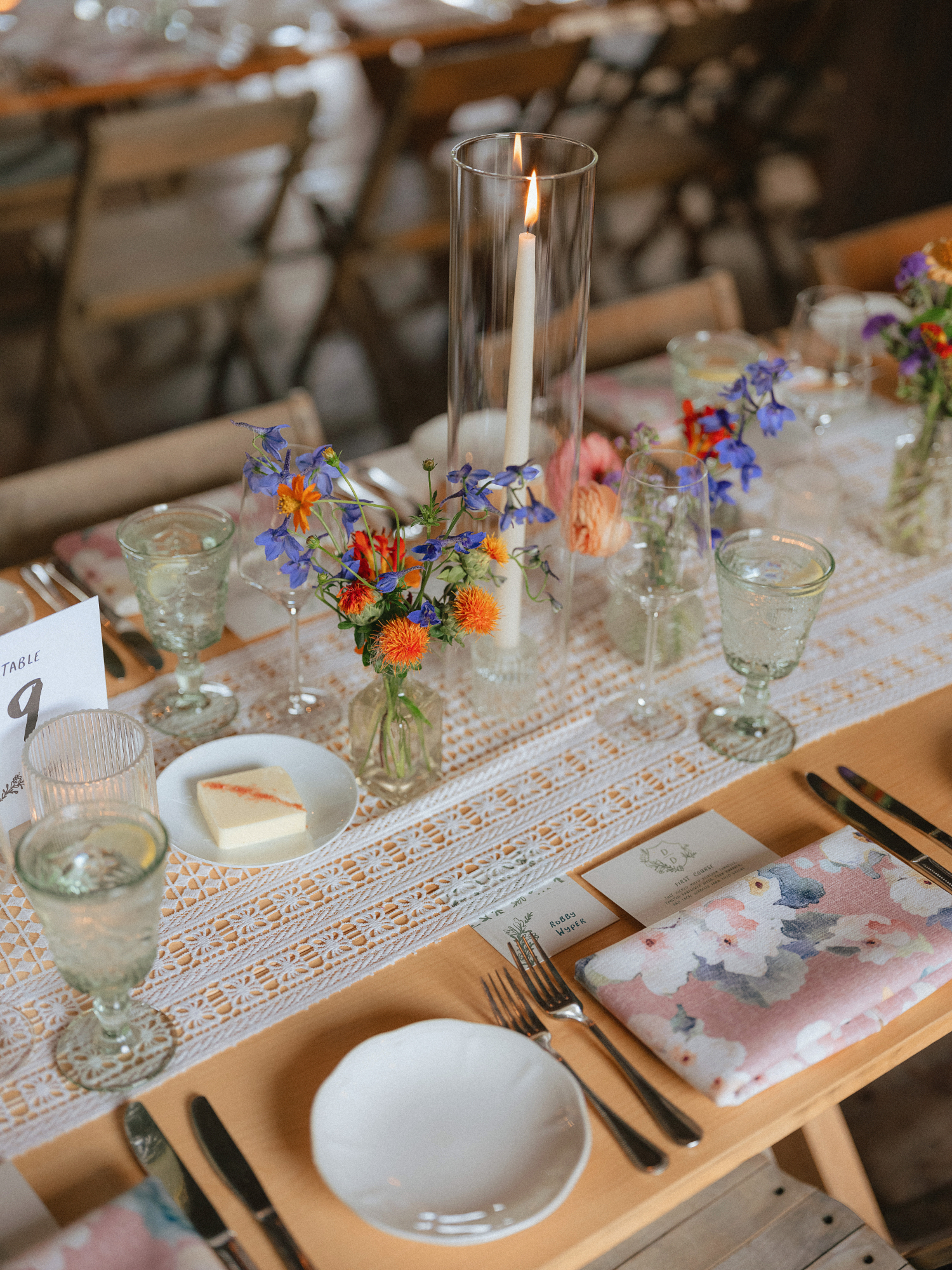 close up shot of wedding reception tablescape details. featuring white plates, and peach, orange, purple, and burgundy blooms placed on white textured table runner and taper candle in hurricane vase