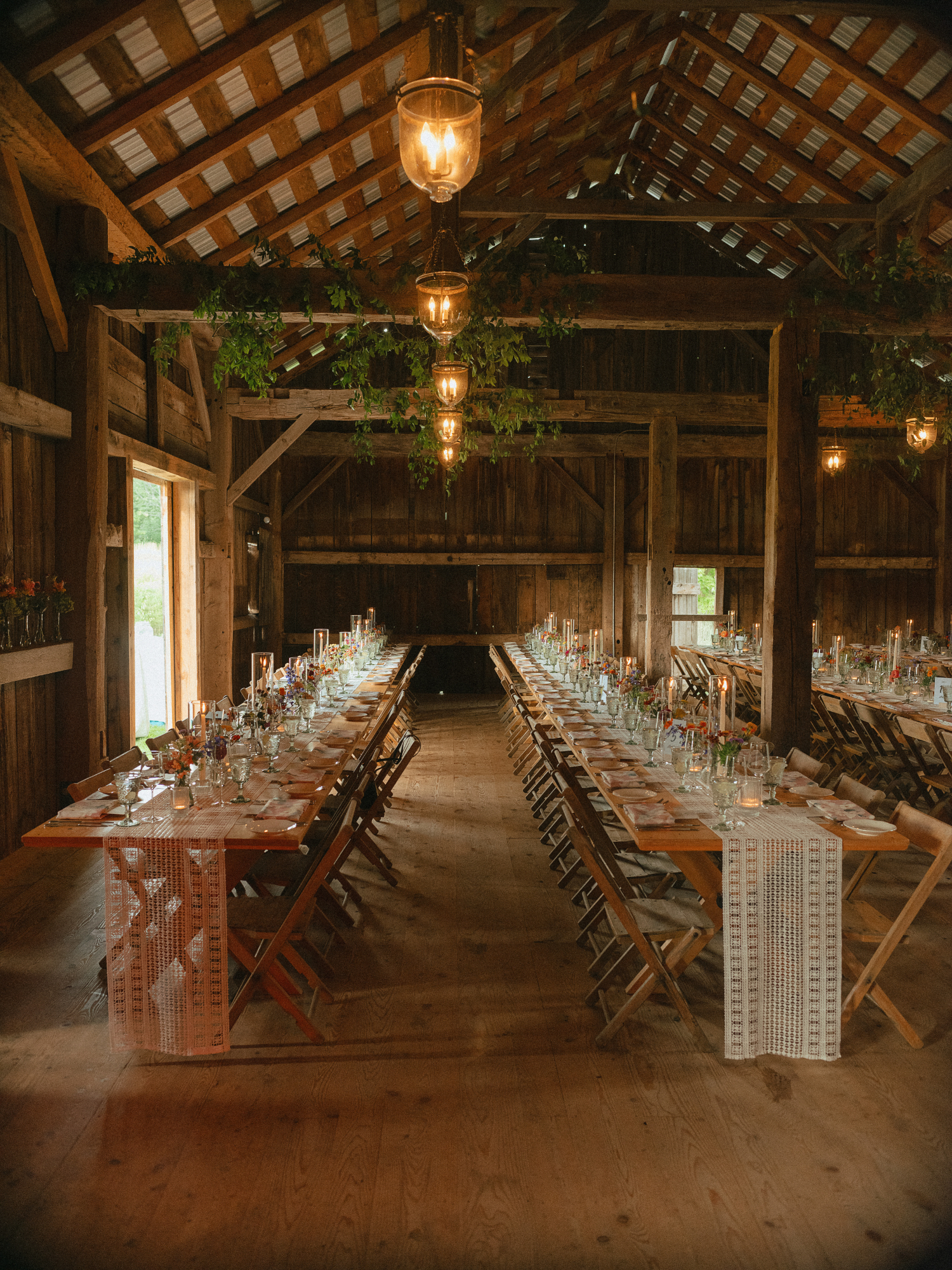 shot of wedding reception tablescapes featuring white plates, and peach, orange, purple, and burgundy blooms placed along the long tables sitting on white textured table runner and taper candle in hurricane vase