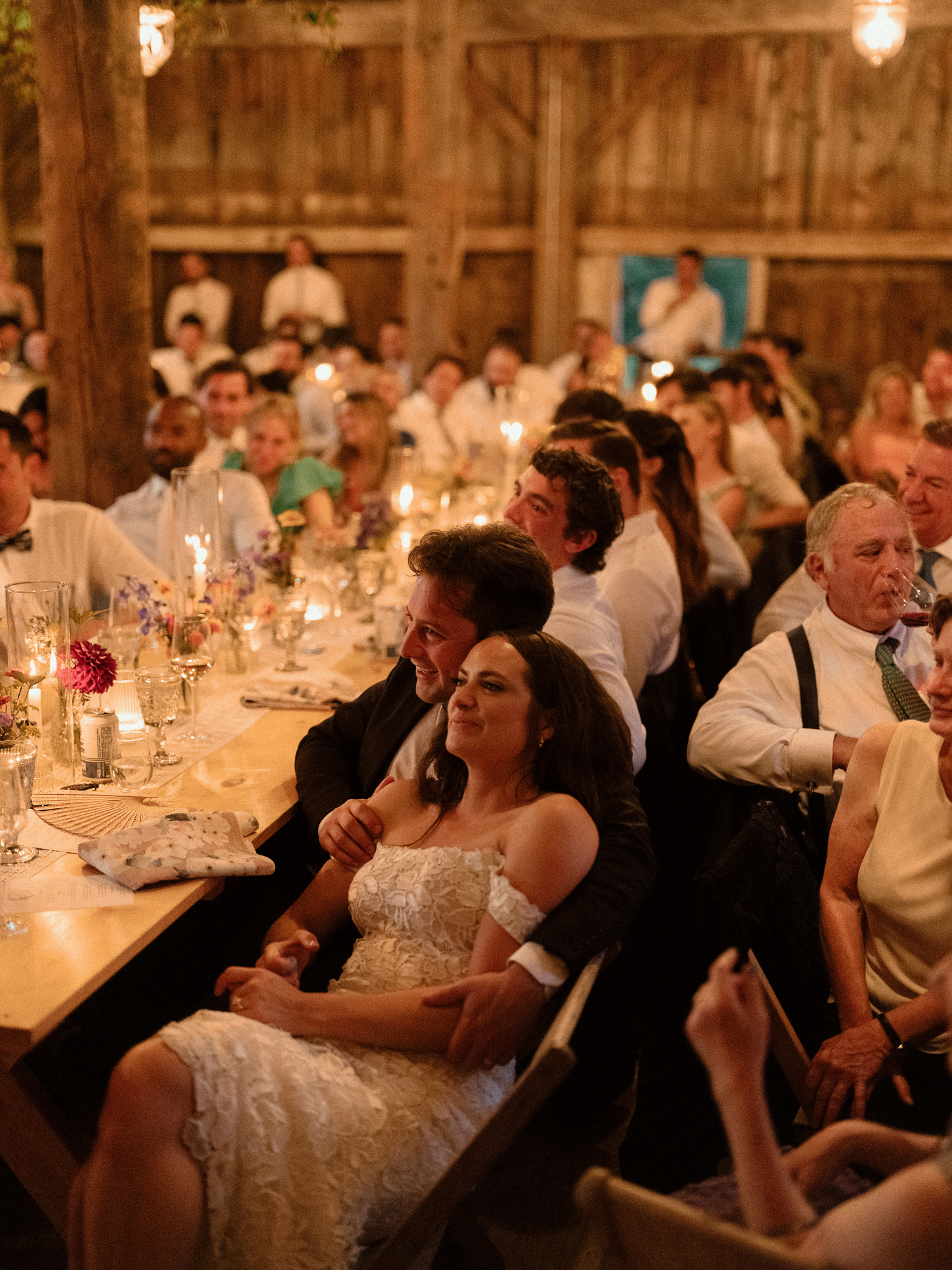 bride and groom sitting together during wedding reception speeches leaning on each other with guests surrounding them