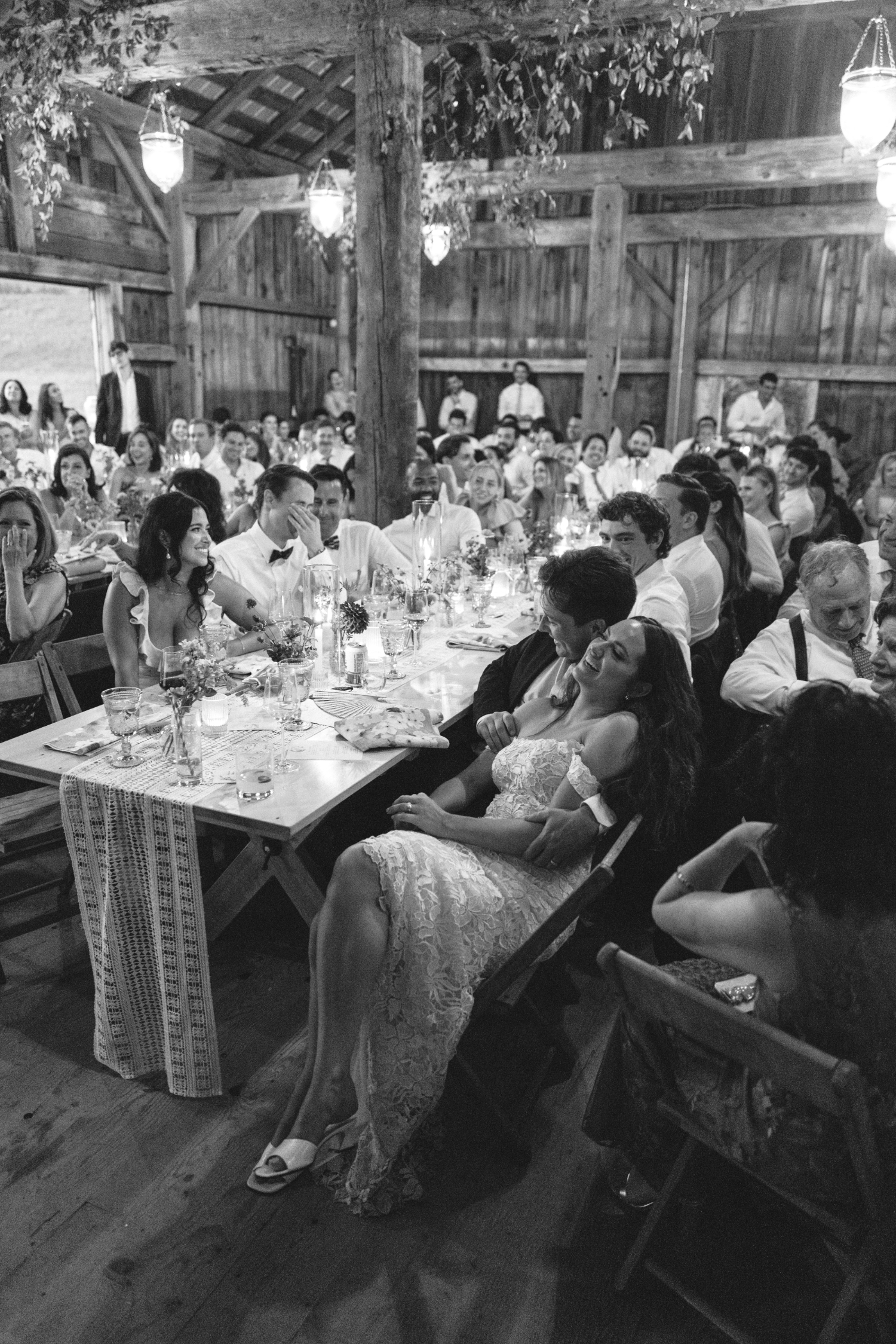 black and white shot of bride and groom sitting together smiling during wedding reception speeches leaning on each other with guests surrounding them