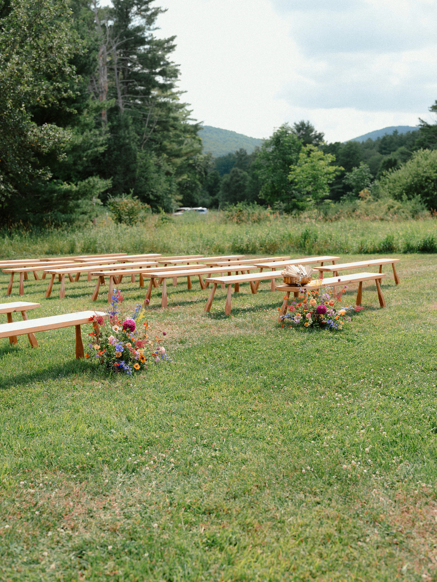 minimalist wooden bench ceremony seating featuring bright aisle arrangements with vibrant pink dahlias, blue delphiniums, orange zinnias, and pops of yellow