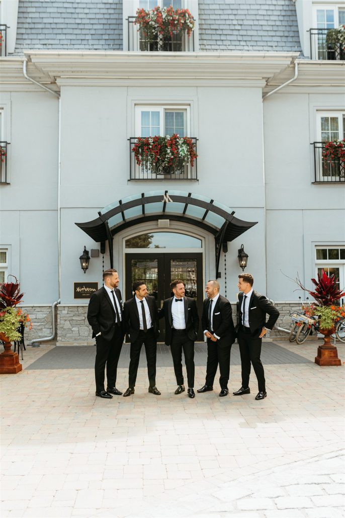 Groom and groomsmen stand confidently in front of a hotel in black tuxedos—transportation and lodging should be factored into the cost of home wedding weekends.