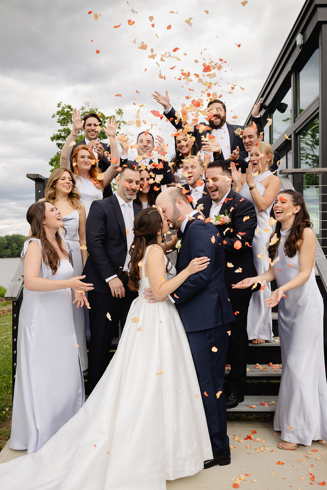 Bride and groom kiss surrounded by their wedding party tossing rose petals, celebrating a joyful moment outdoors. Details handled in wedding planner vs venue coordinator roles.
