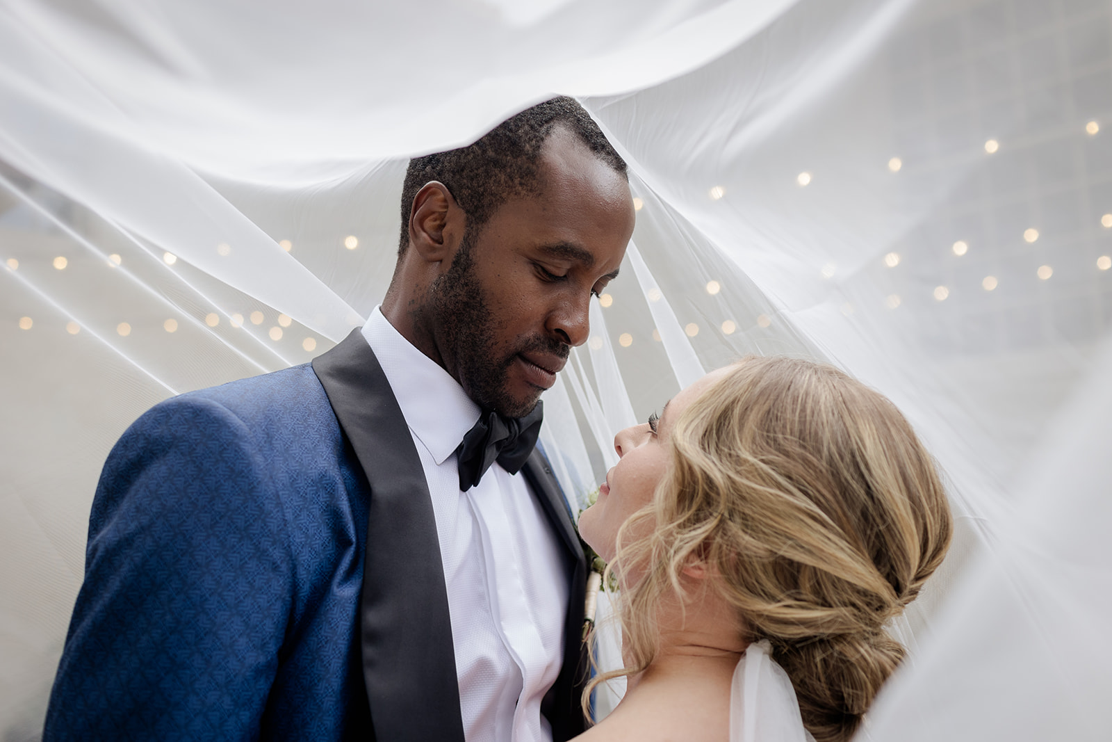 Bride and groom share an intimate moment under a veil with twinkle lights in the background—timeless and romantic.