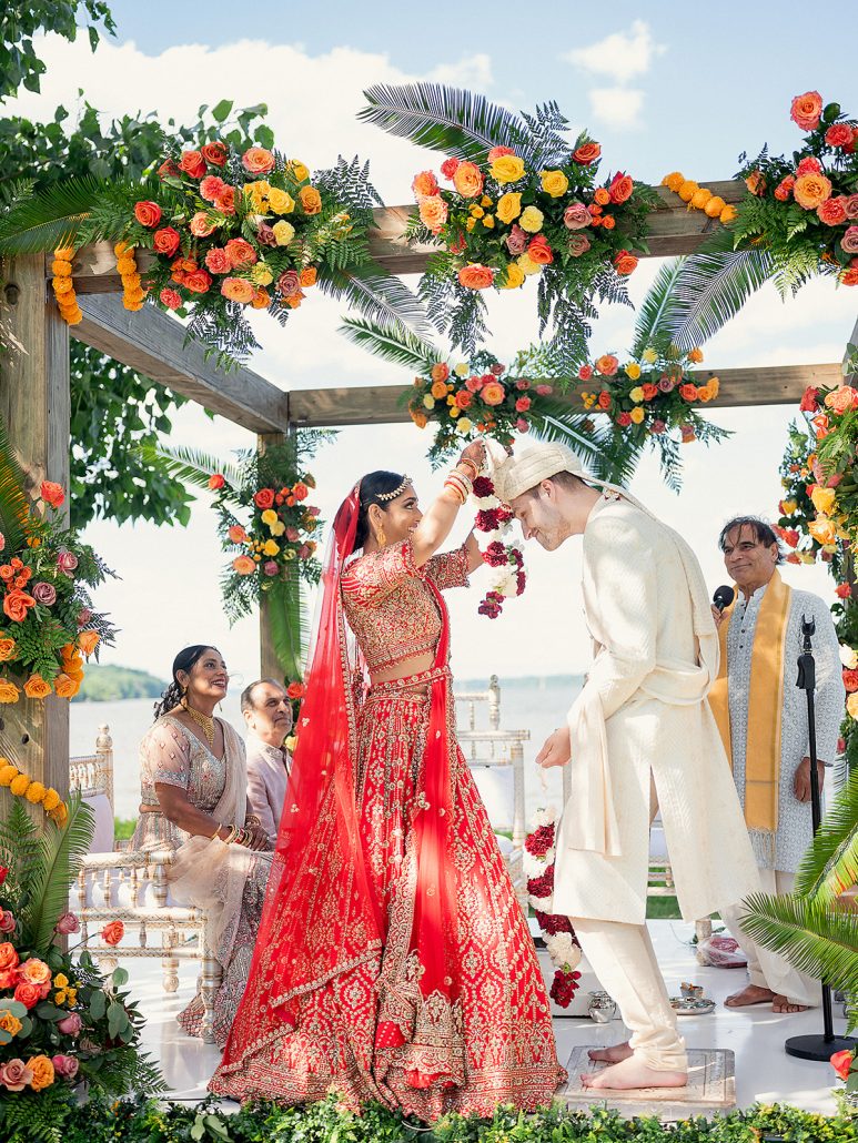 Bride placing floral garland on groom under a vibrant floral mandap by the Hudson River.