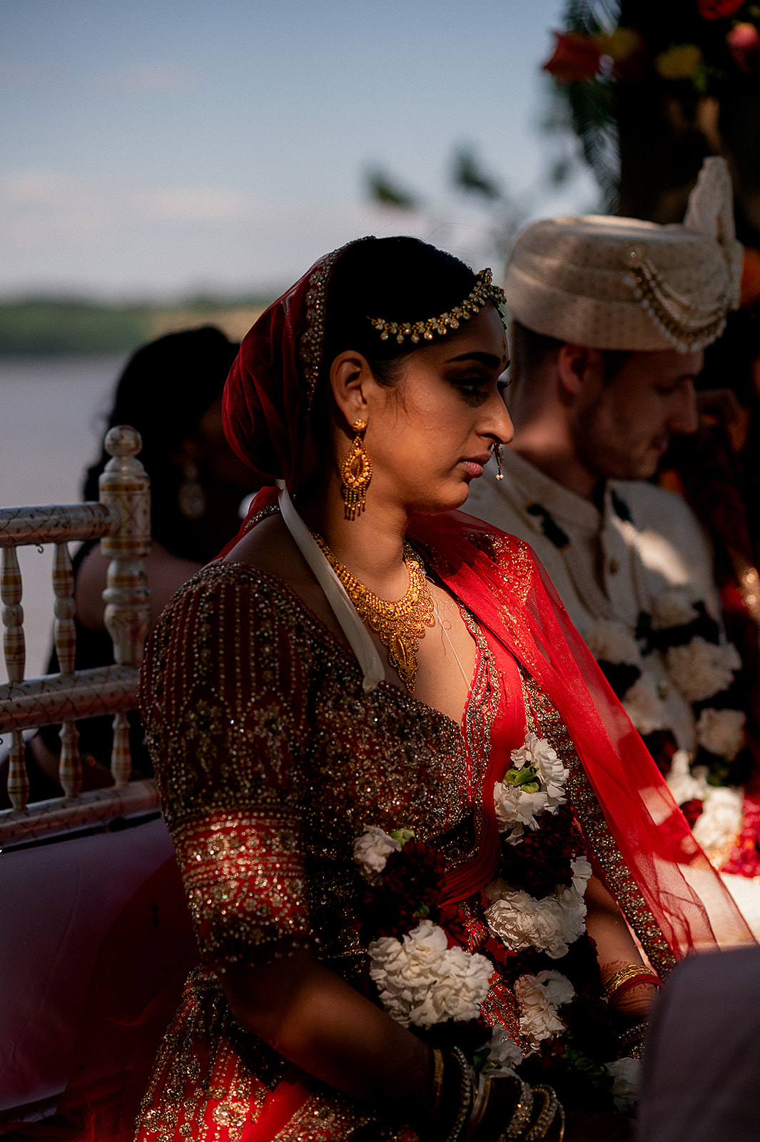 Bride seated at mandap with traditional jewelry and floral garland, bathed in golden light.