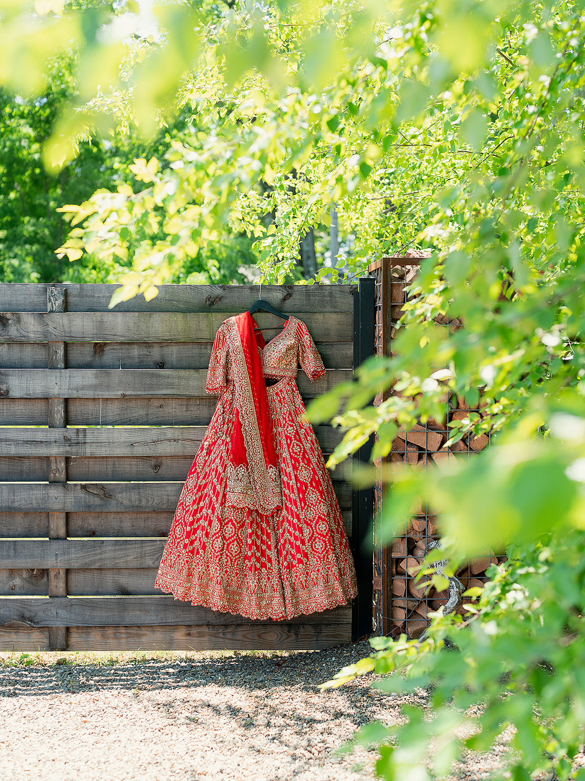 Full view of a red Indian bridal lehenga hanging on a rustic wooden fence surrounded by greenery.