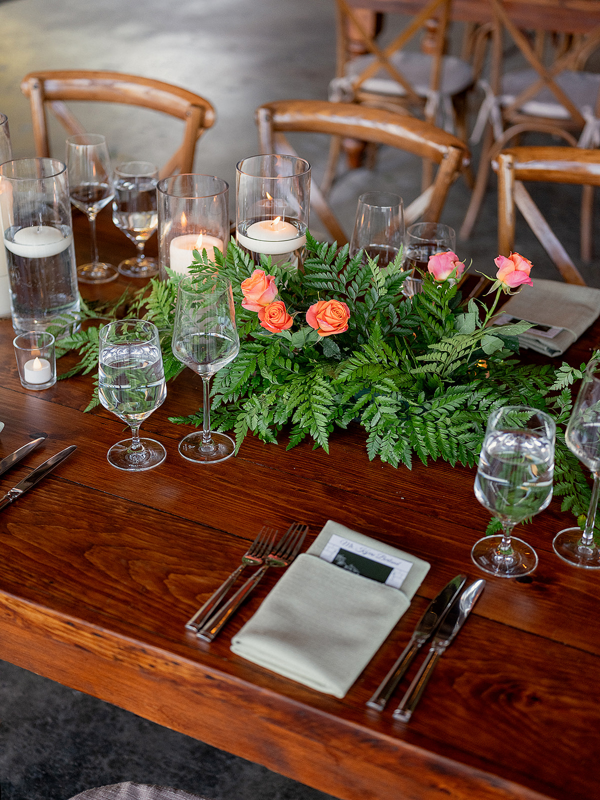 Elegant wedding tablescape with lush greenery, coral roses, floating candles, and fine glassware on a rich wooden table.