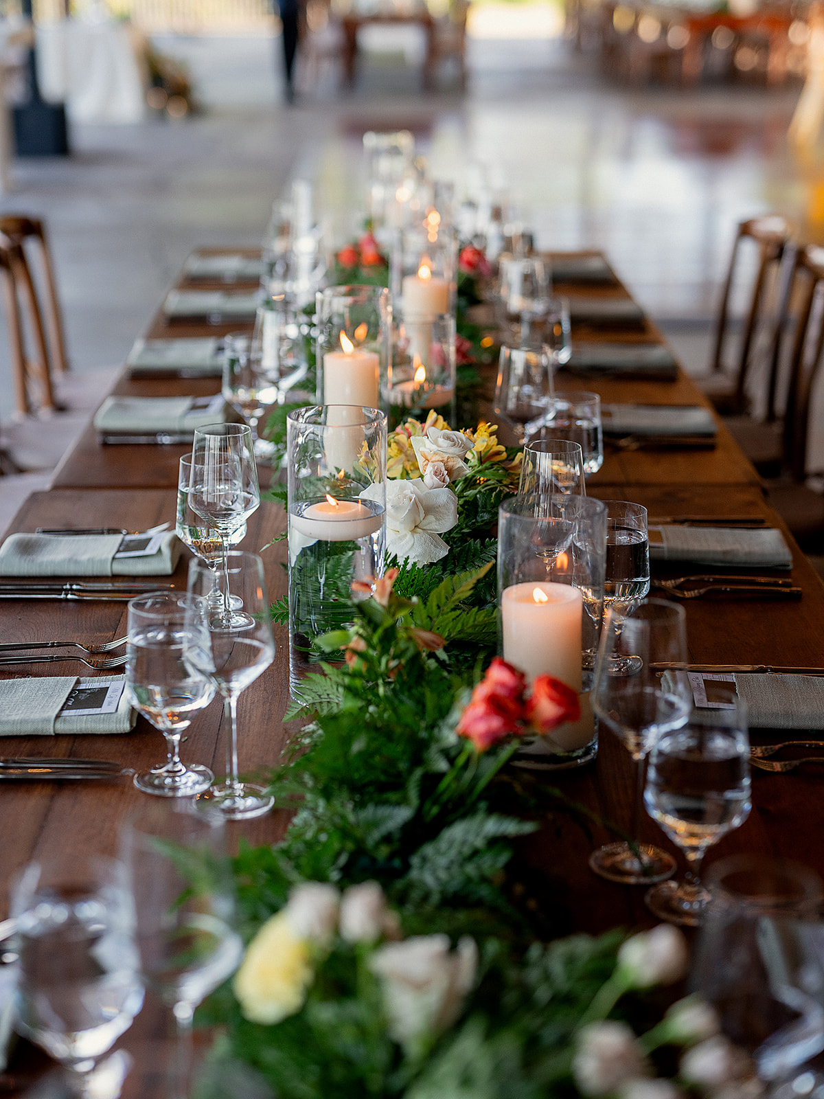 Long reception table featuring vibrant florals, pillar candles in glass vases, and a mix of blush and coral blooms arranged with ferns.