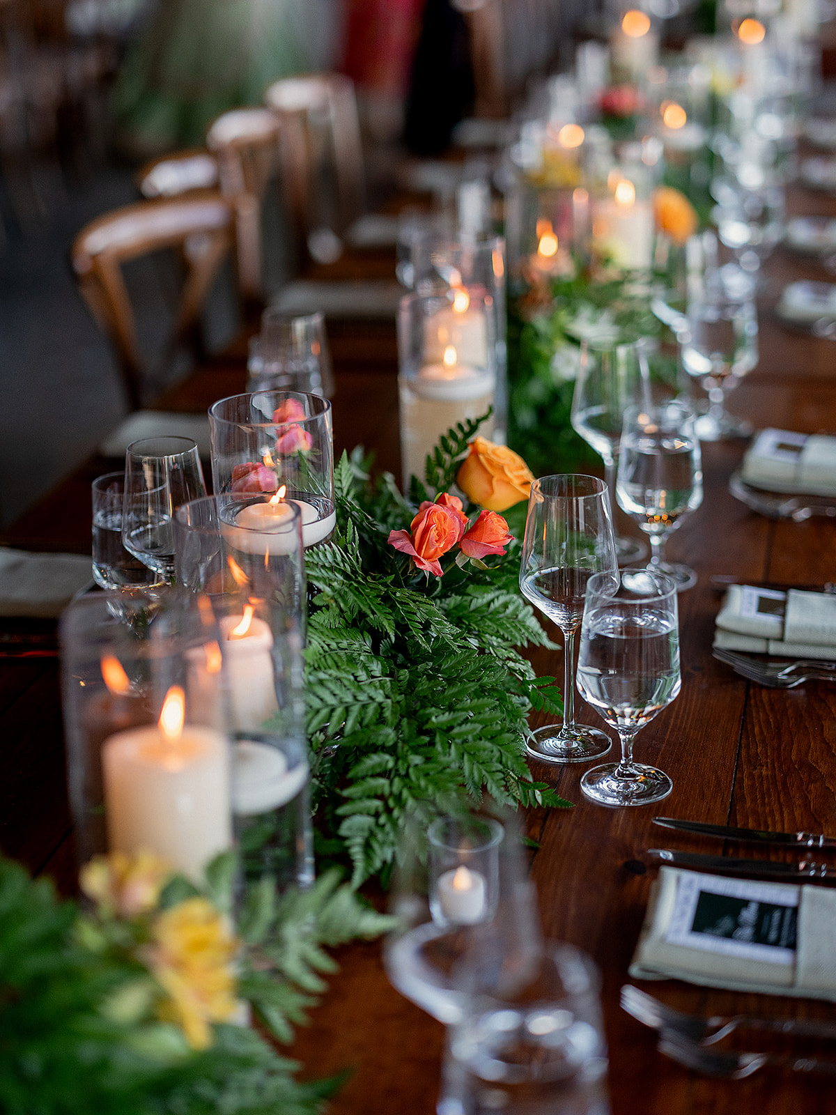 Long reception table featuring vibrant florals, pillar candles in glass vases, and a mix of blush and coral blooms arranged with ferns.