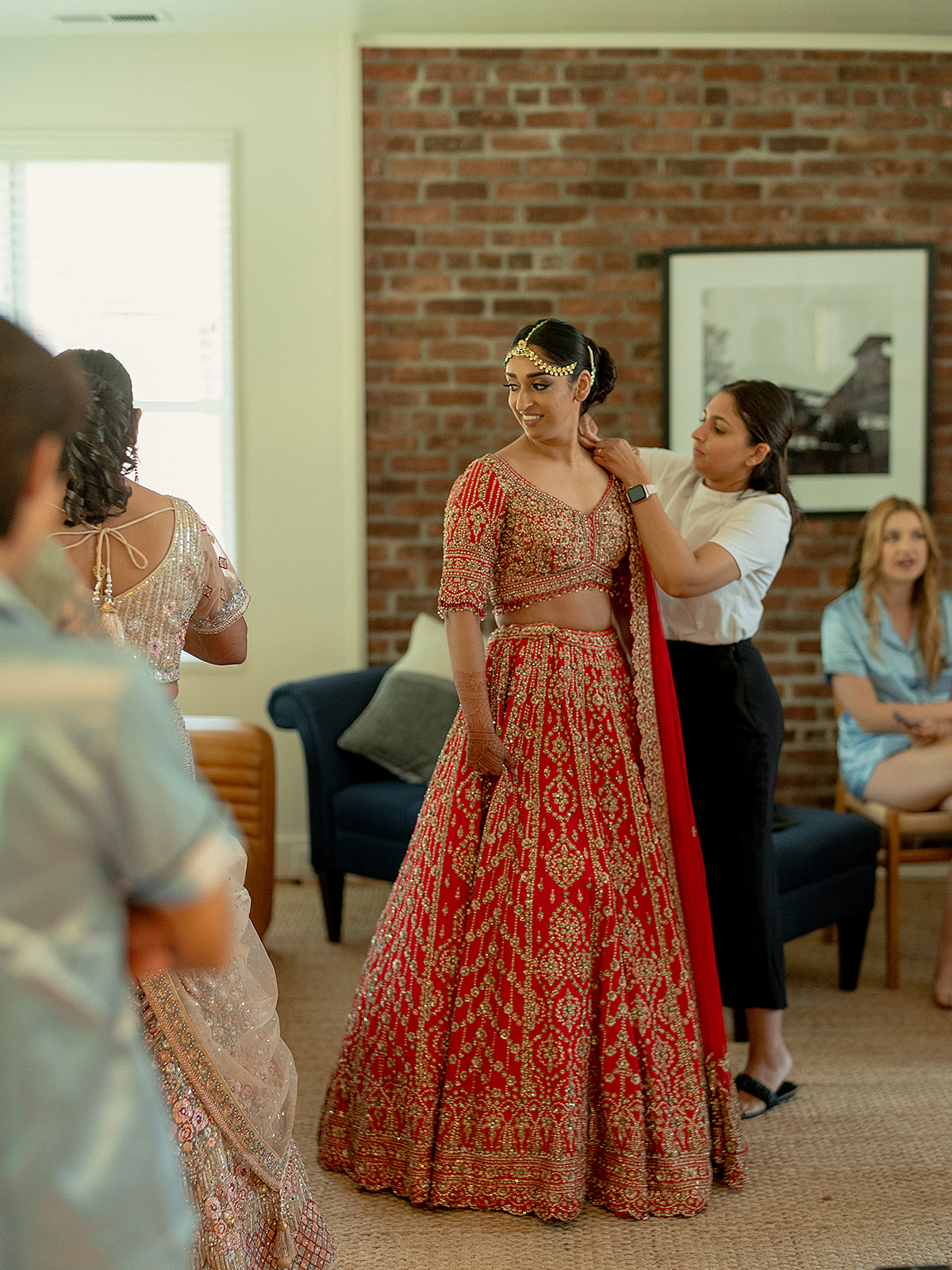 Bride getting ready for her Indian wedding in a red lehenga, assisted by her glam team indoors.