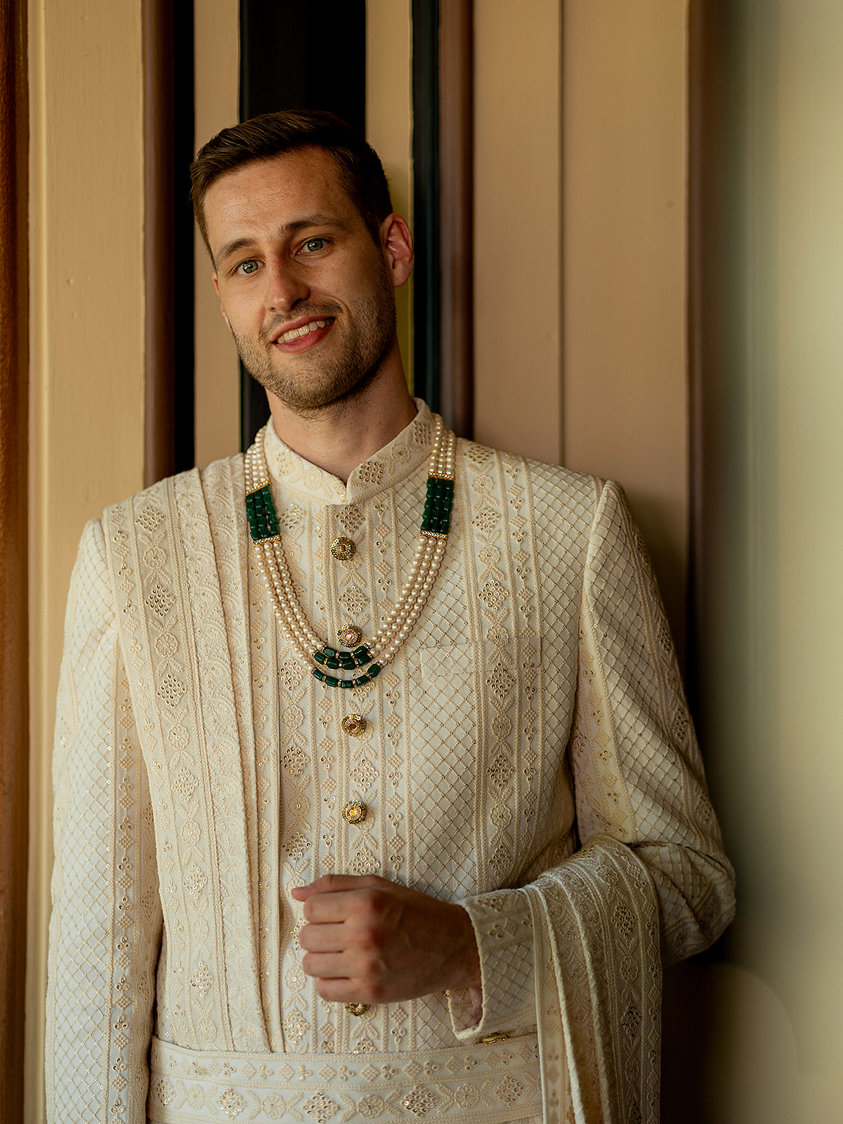 Groom in ivory sherwani and pearl necklace, posing for a portrait before the Indian wedding ceremony.