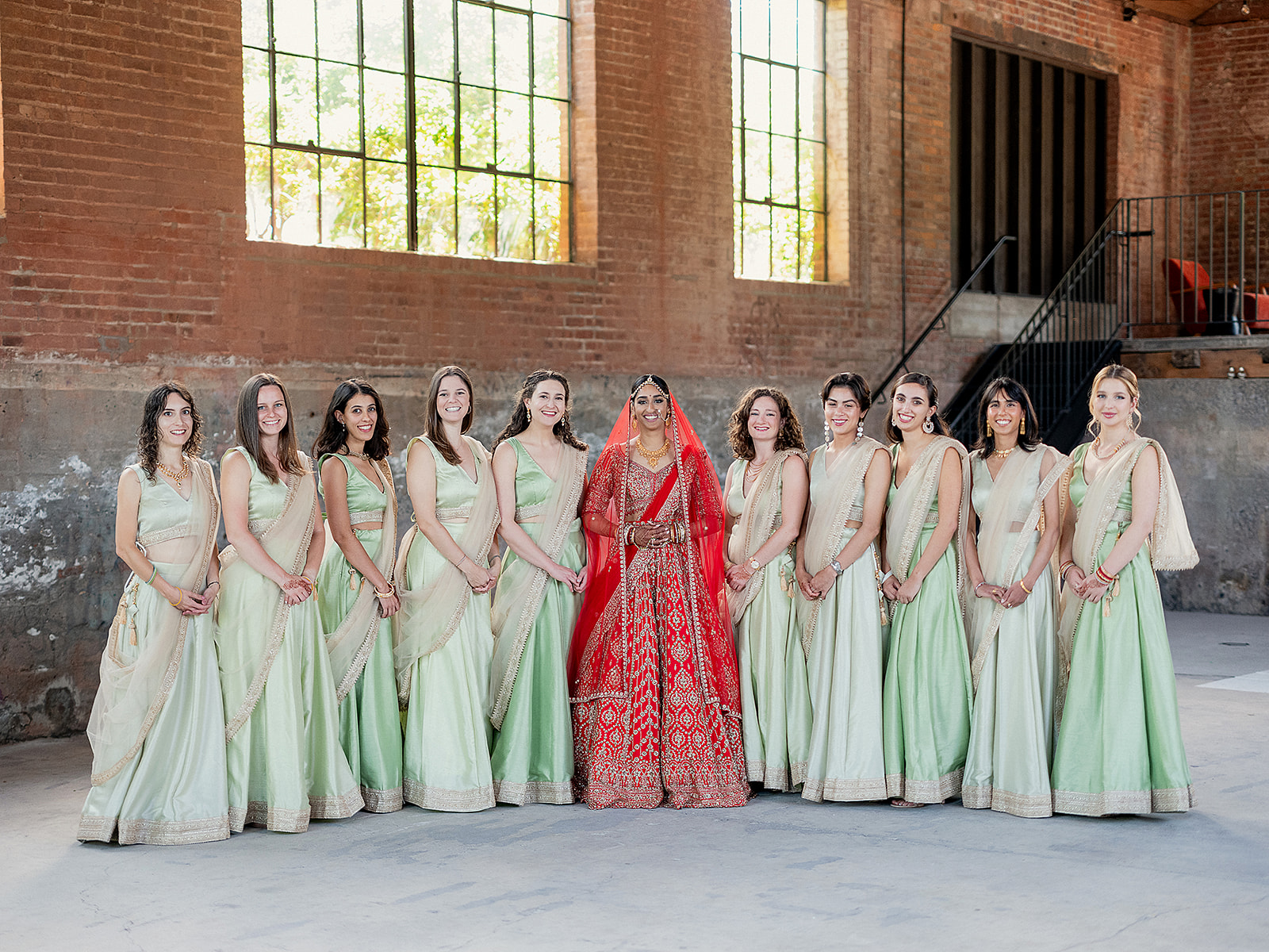 Bride standing with her bridesmaids dressed in sage lehengas in a brick-lined wedding venue.