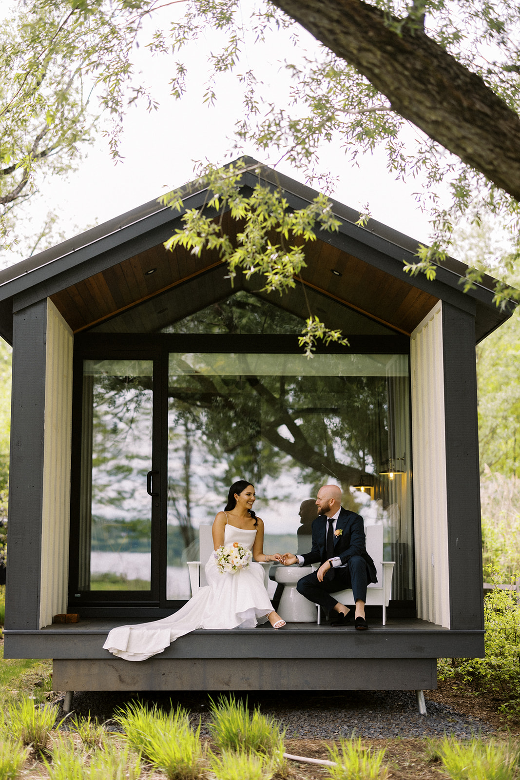 Bride and groom sit on the porch of a modern cabin, holding hands and smiling during their intimate wedding weekend.