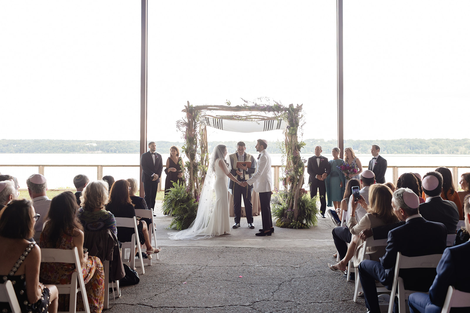Couple exchanges vows under a chuppah with river views behind them, surrounded by loved ones in a waterfront wedding ceremony.