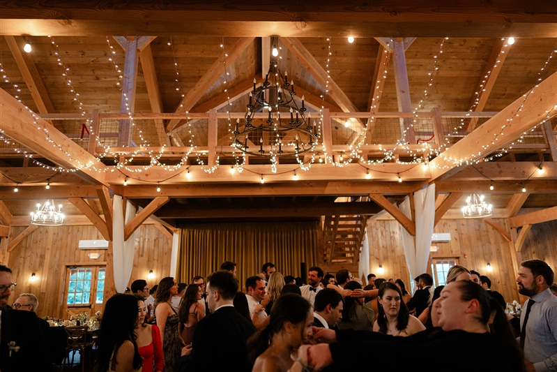 Guests dancing under twinkle lights and wood-beamed ceilings during a barn wedding reception.