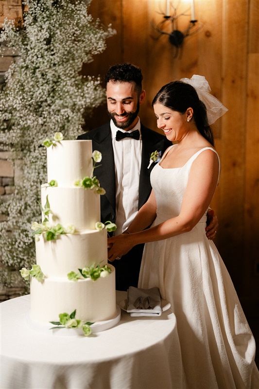 Bride and groom cutting a modern white wedding cake decorated with green florals inside a warm, wood-paneled reception space at a Hudson Valley wedding venue with accommodations.