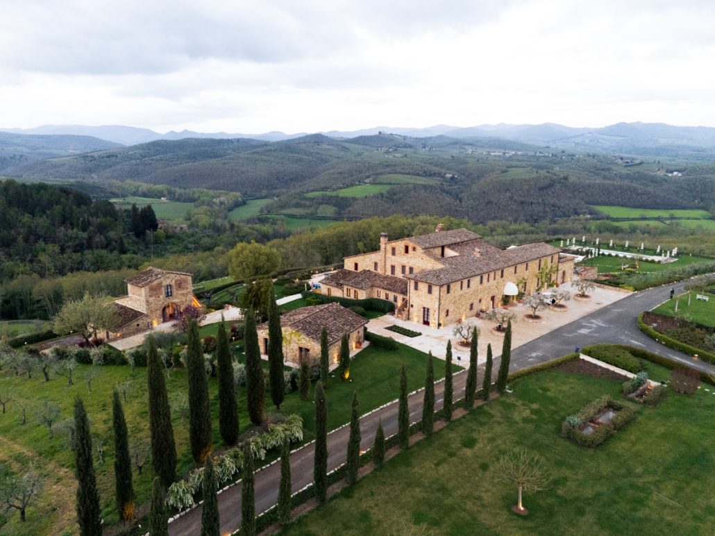 Panoramic shot of wedding venue in Tuscany with a stone villa, chapel, and cypress-lined driveway surrounded by vineyards.