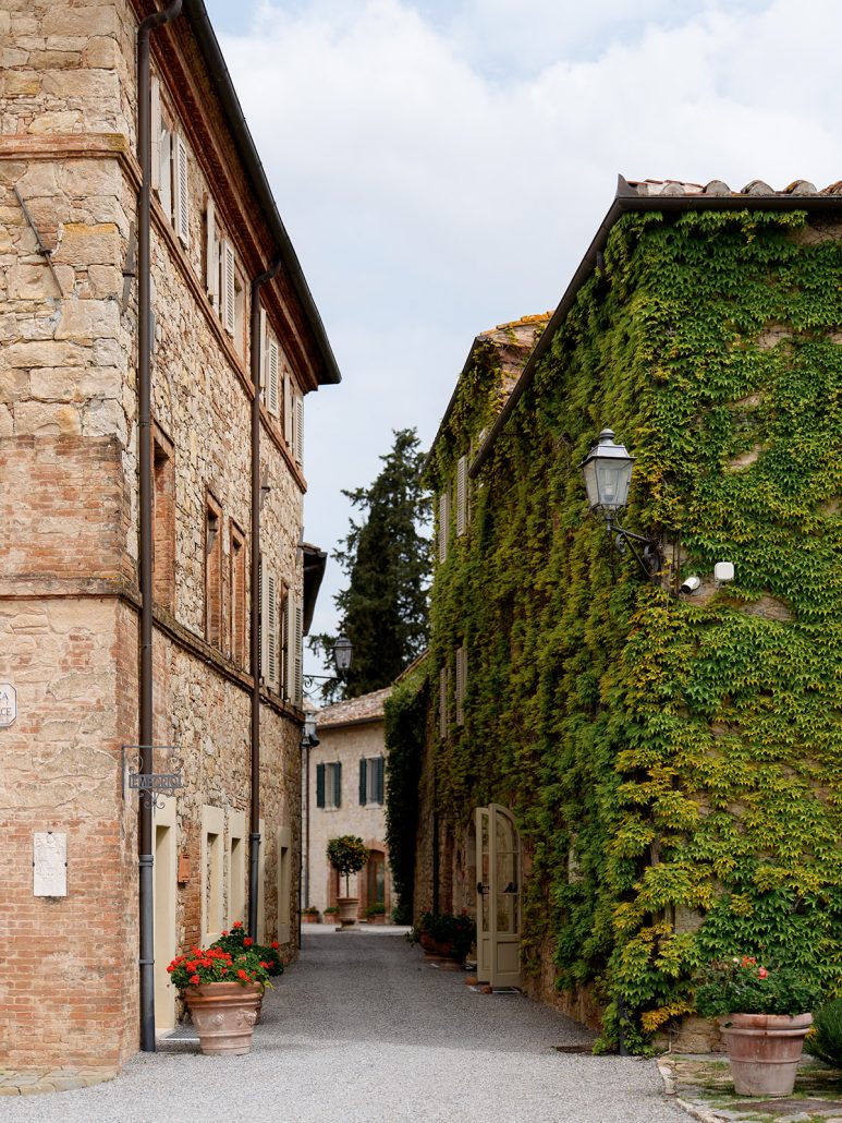 Narrow cobblestone alley in Borgo San Felice Resort, Tuscany, with ivy-covered stone walls, wooden shutters, and terracotta pots of red flowers.