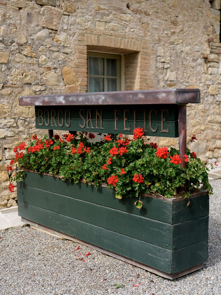 Wooden planter box with vibrant red geraniums and “Borgo San Felice” sign, set against rustic stone walls of this Tuscany wedding venue.