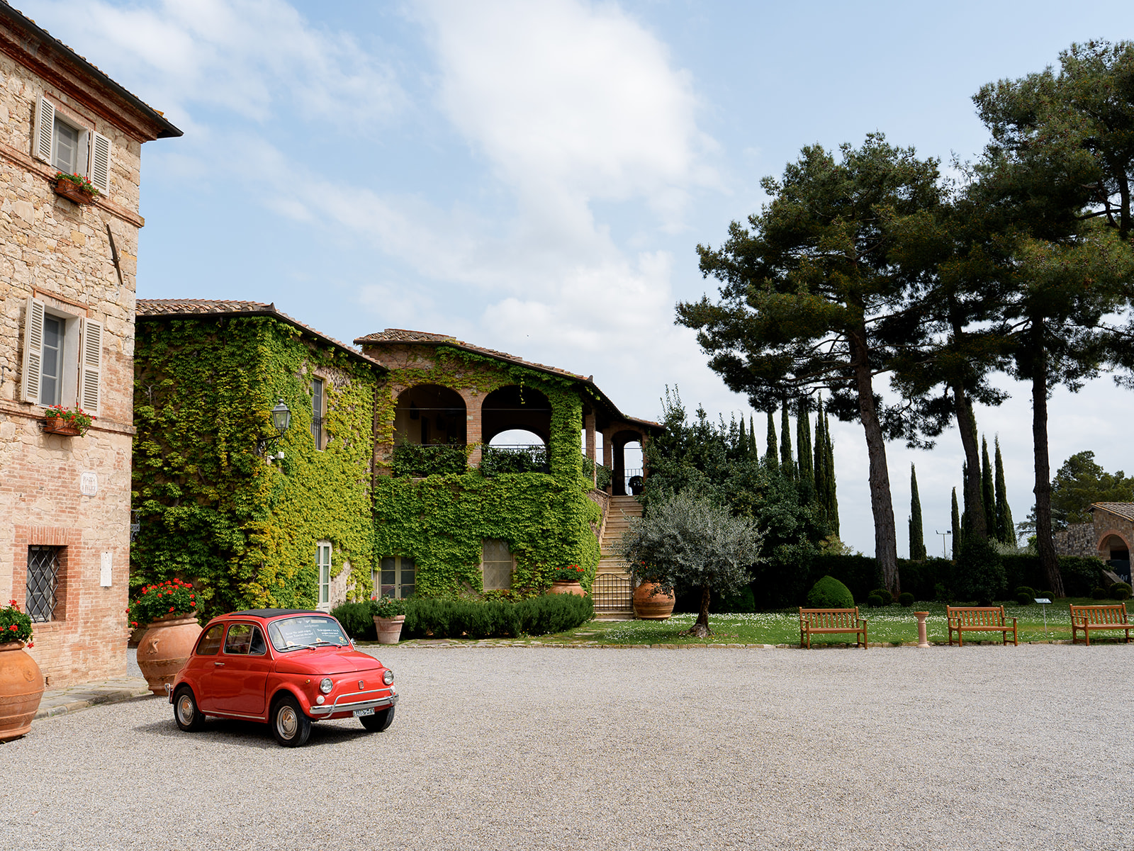 Courtyard at Borgo San Felice Resort with ivy-covered walls, tall cypress trees, terracotta pots, and a vintage red Fiat 500.