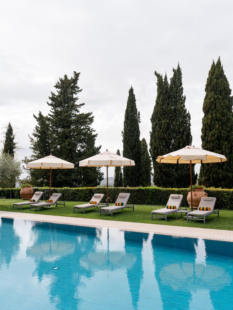 Swimming pool at Borgo San Felice Resort, Tuscany, lined with lounge chairs, parasols, and terracotta pots against cypress trees.