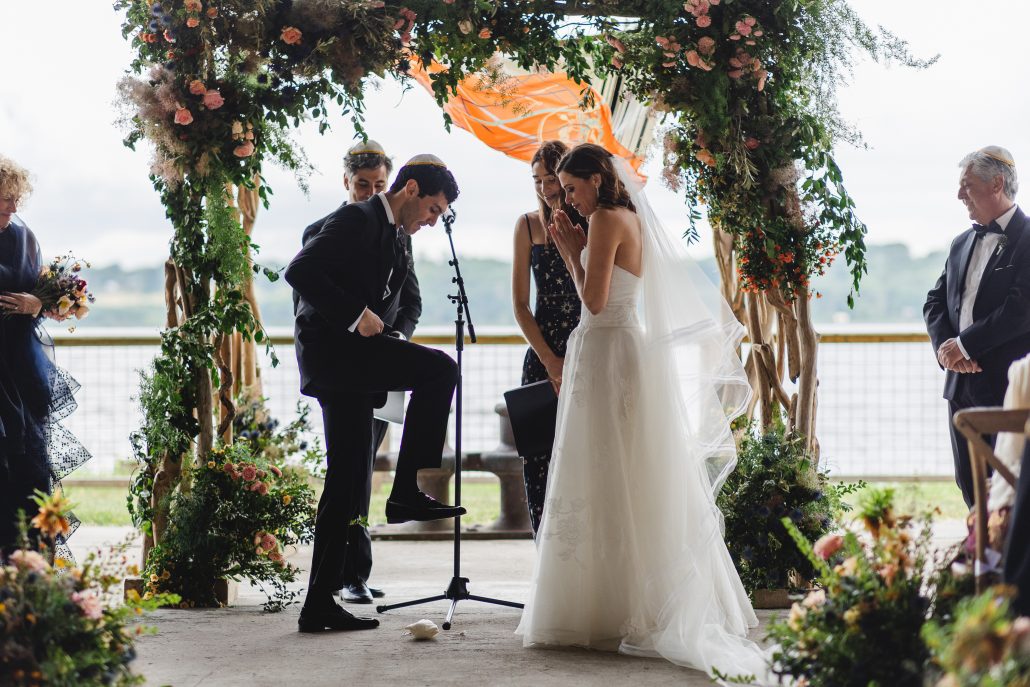 A groom breaks the glass under a floral chuppah during a lakeside Jewish wedding—showcasing tradition within modern 2026 wedding trends.