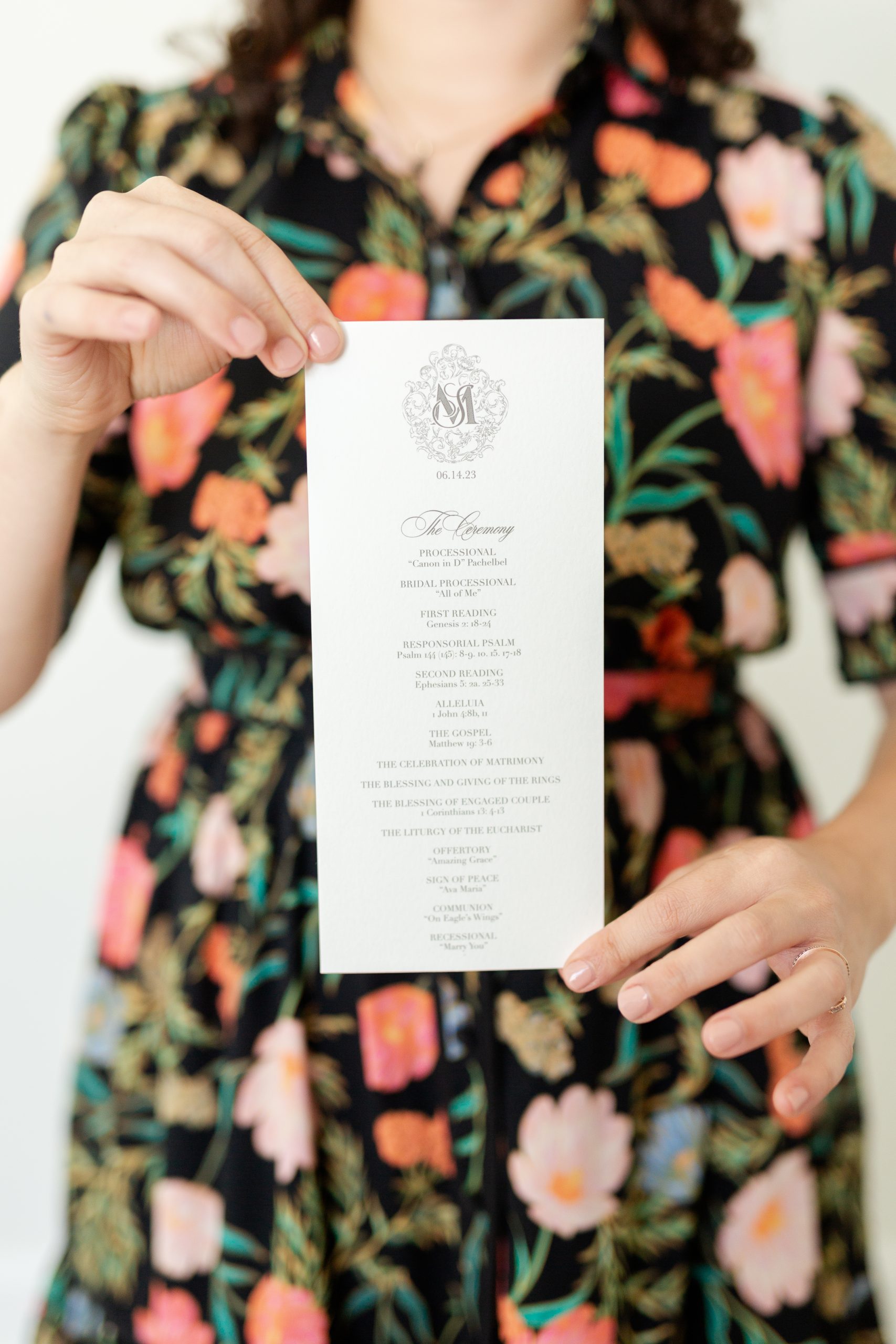 A woman in a floral dress holding a vertically designed wedding ceremony program featuring elegant script and monogram detailing. mailing wedding invitations