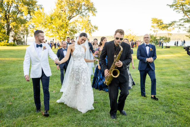 Bride and groom walking hand in hand behind brass band musicians during their summer Hudson River wedding