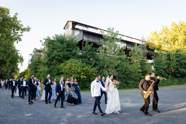 Bride and groom smiling as a saxophonist leads their wedding procession at Hutton Brickyards