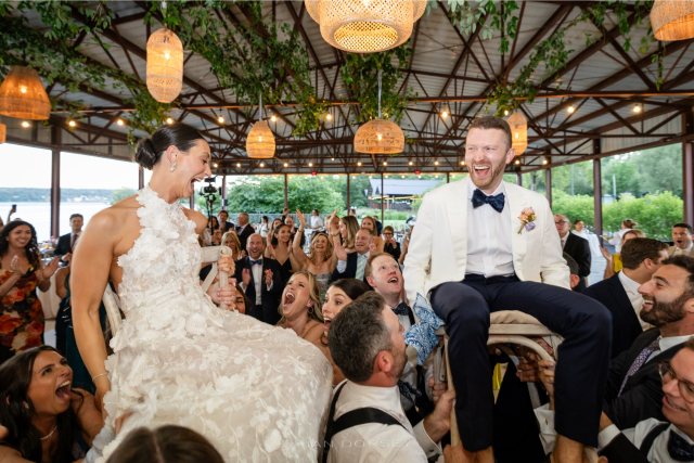 The couple laughing as they are lifted on chairs during the hora, surrounded by cheering guests and rustic chandeliers.