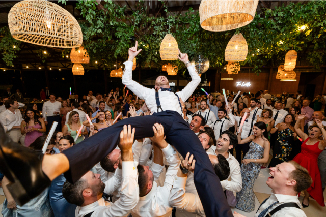 The groom being lifted high into the air on the dance floor, smiling and holding glow sticks as guests cheer beneath hanging lights.