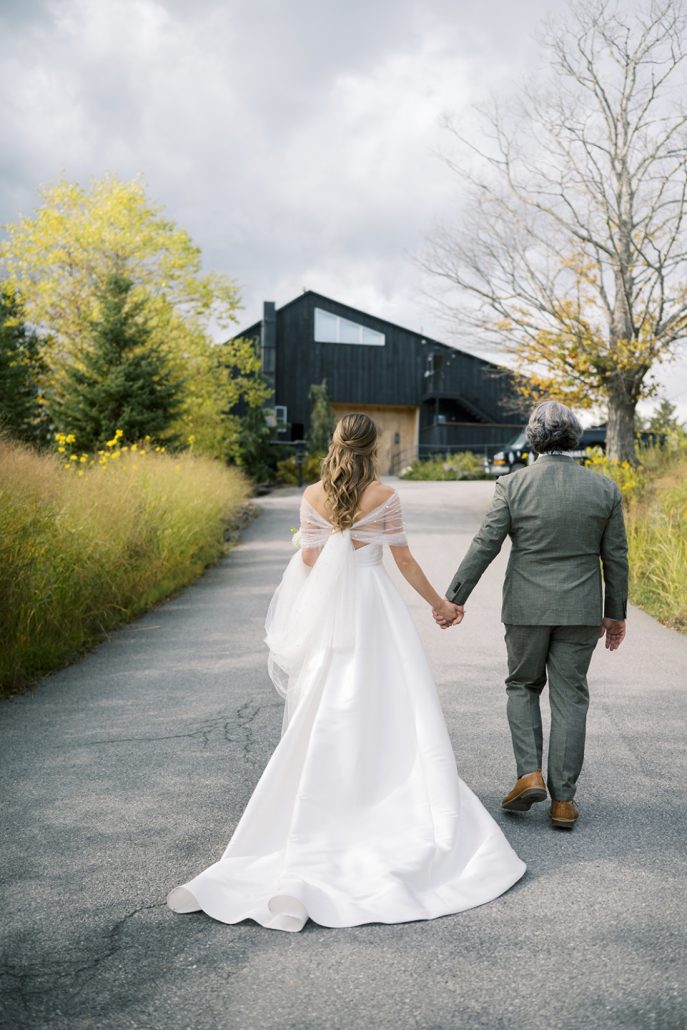 Bride and groom walking hand in hand toward the lodge along a tree-lined path.