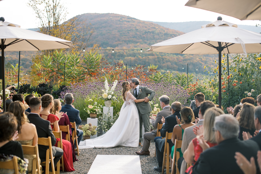 Couple shares a kiss during an outdoor ceremony framed by fall mountain views at Scribner’s Catskill Lodge wedding.