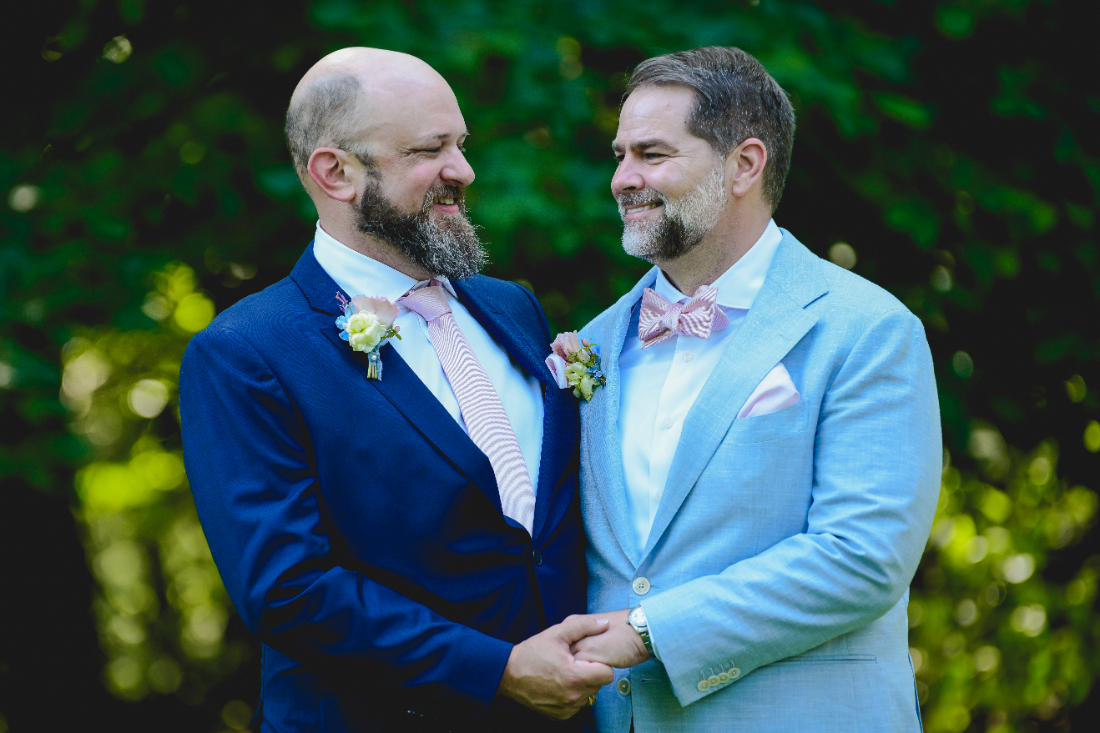 Grooms hold hands and smile at each other during their buttermilk falls inn wedding, standing outdoors against lush green Hudson Valley scenery.