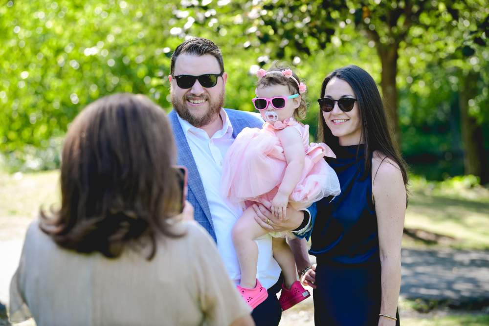 A family smiles for a photo with their toddler in a pink dress and sunglasses during a buttermilk falls inn wedding.
