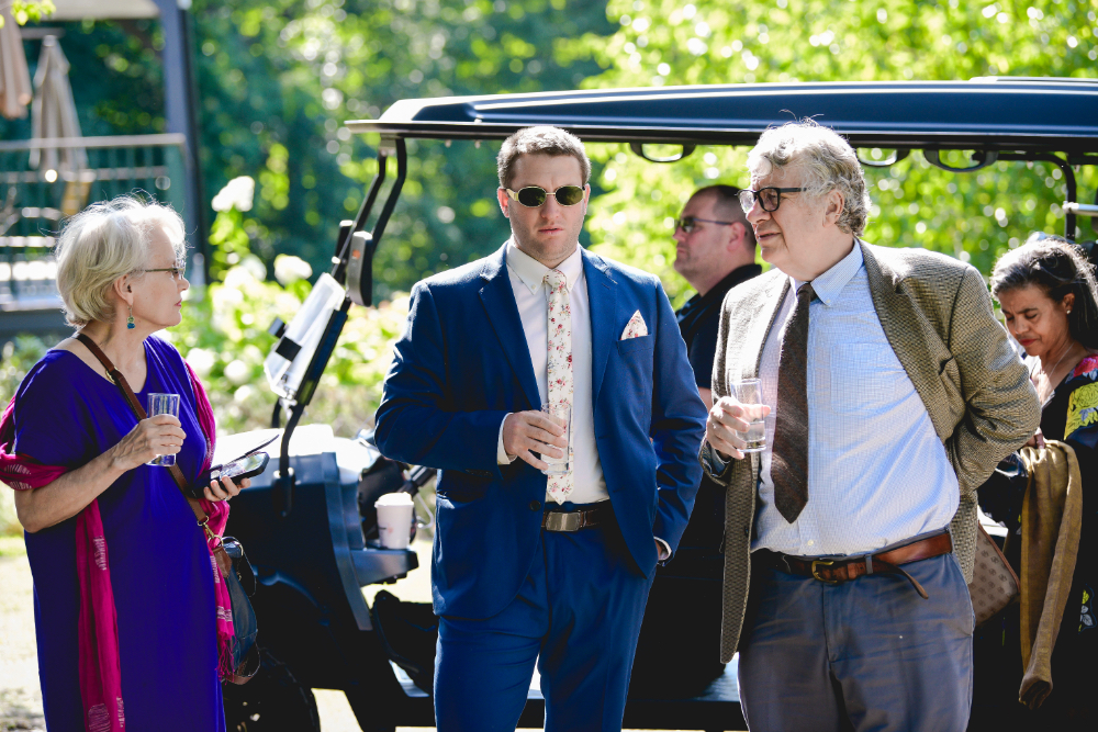 Wedding guests in suits and dresses enjoy drinks and conversation outdoors during a wedding.