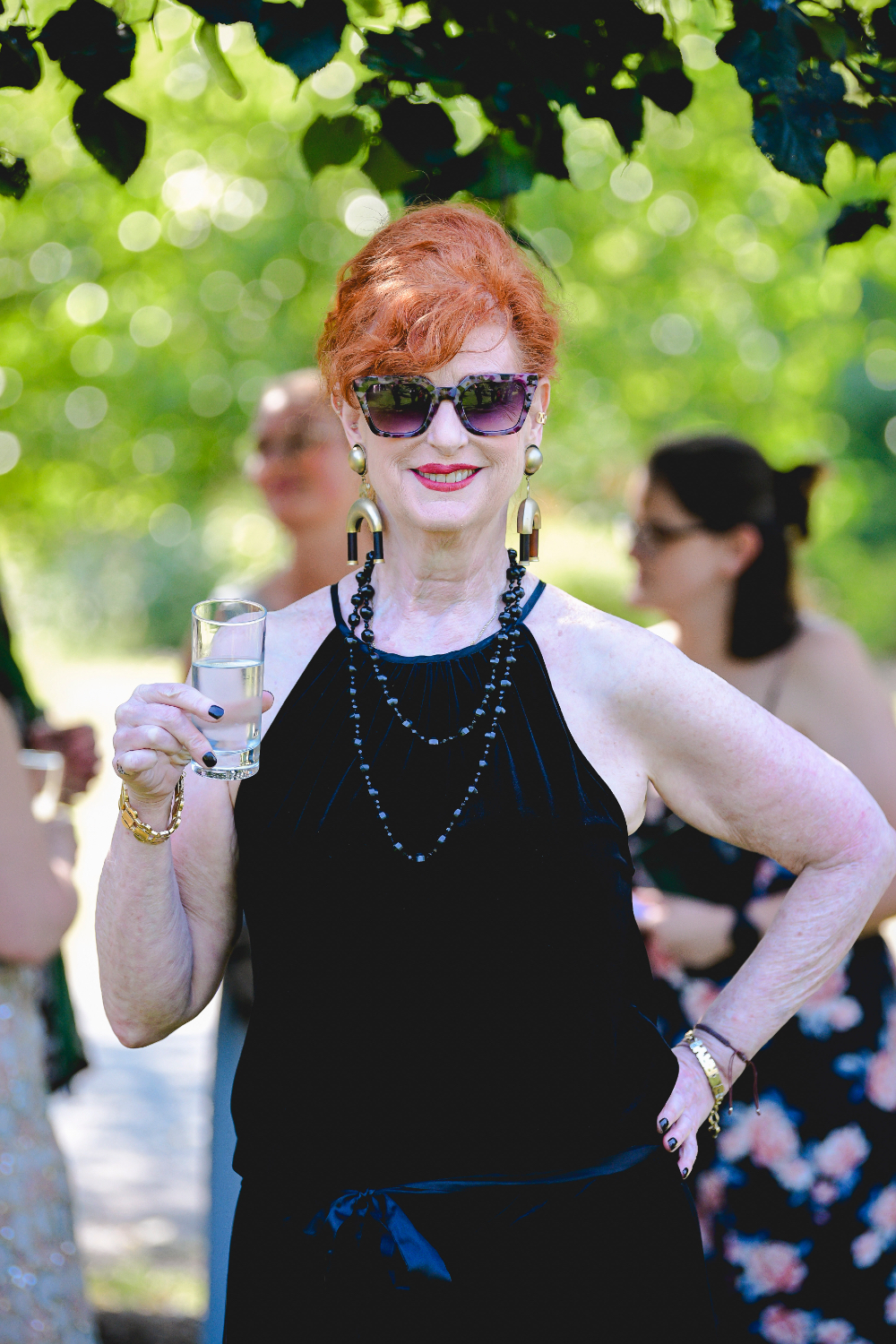 A stylish guest holding a drink and smiling beneath the trees during a buttermilk falls inn wedding.