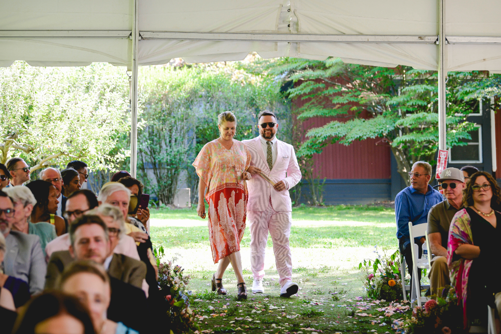 Two guests walk arm in arm down the flower-lined aisle under the ceremony tent at a buttermilk falls inn wedding.