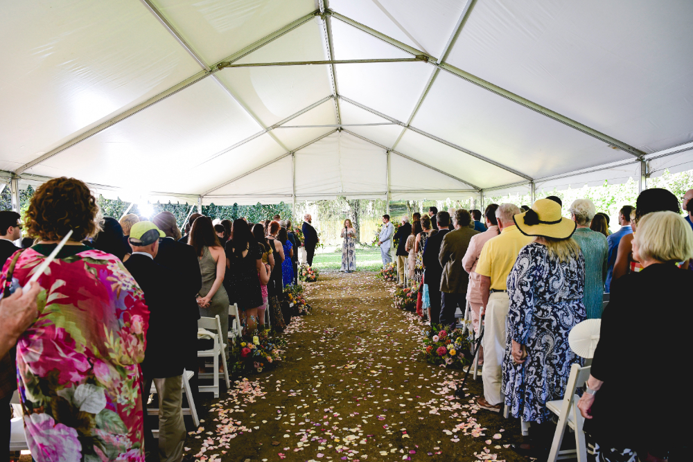 A wide view of a Buttermilk Falls Inn wedding ceremony inside a white tent, with guests standing along a petal-covered aisle as the couple stands at the altar.
