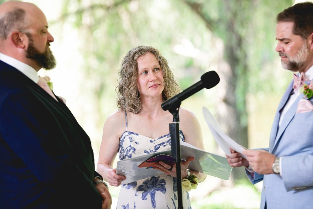 The officiant smiles warmly while leading the vows during a Buttermilk Falls Inn wedding ceremony, with the couple standing on each side amid soft natural light and garden scenery.