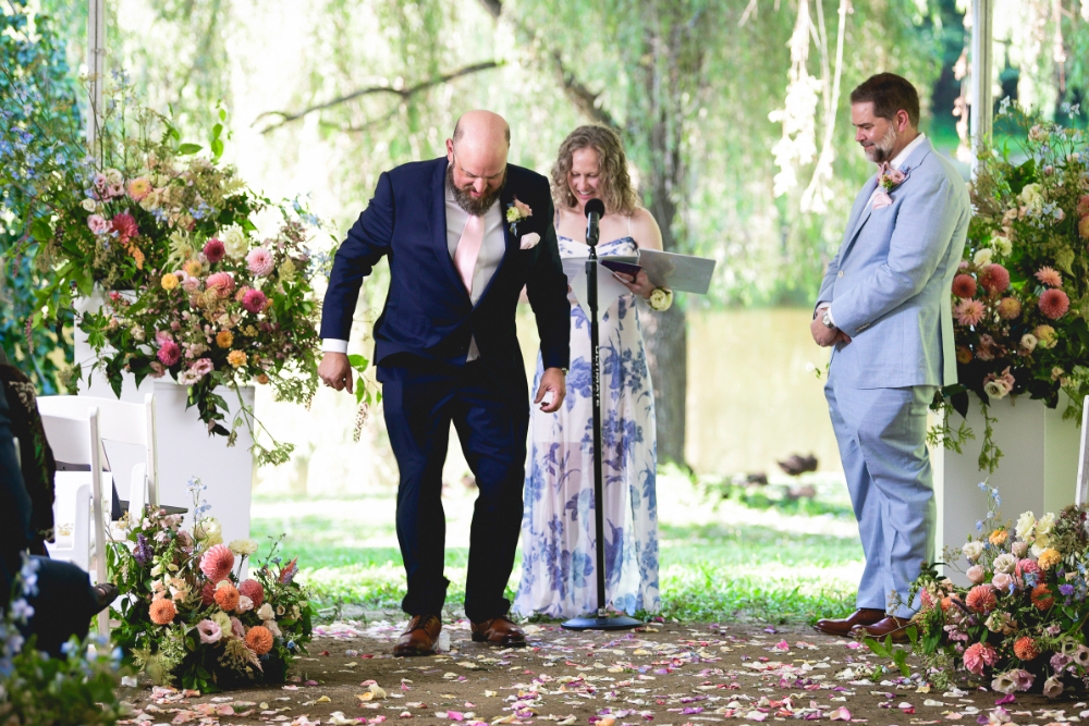 A joyful moment at a Buttermilk Falls Inn wedding as the grooms walk hand in hand down the aisle under a white tent, surrounded by cheering guests and floating bubbles.