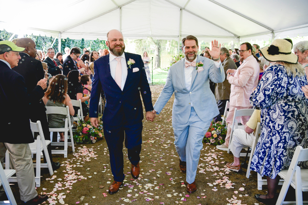 A joyful moment at a Buttermilk Falls Inn wedding as the grooms walk hand in hand down the aisle under a white tent, surrounded by cheering guests and floating bubbles.