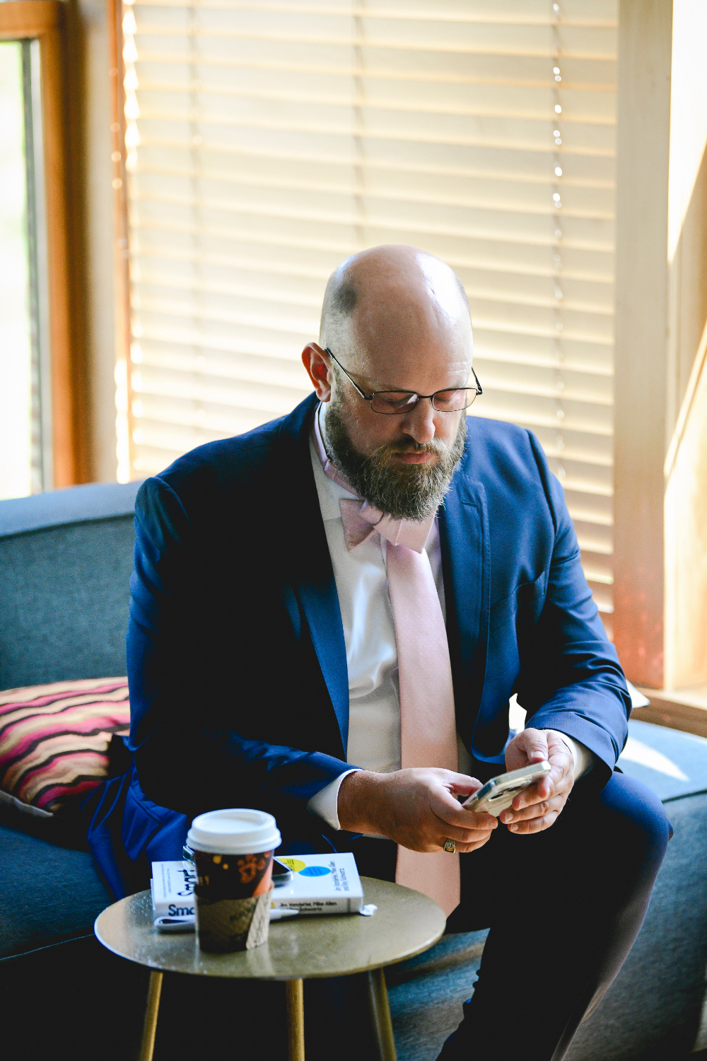 A groom in a navy suit sits by a window checking his phone on the morning of his Buttermilk Falls Inn wedding.