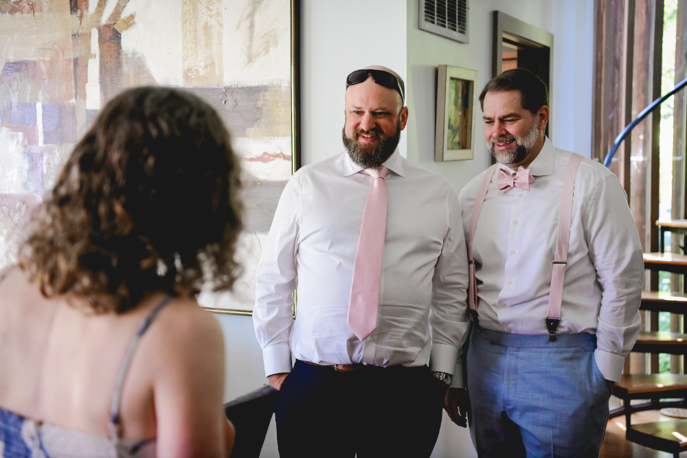 Two grooms in matching white shirts and blush accessories share a light moment with a friend during their Buttermilk Falls Inn wedding morning preparations.