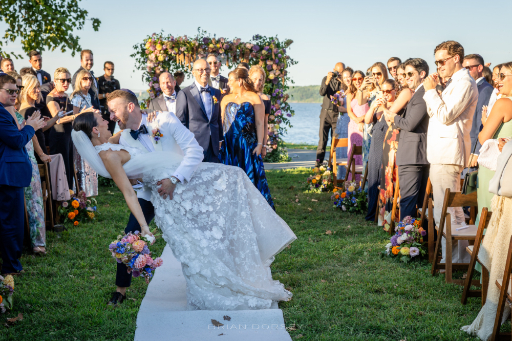 Newlyweds share a joyful dip kiss at their outdoor ceremony, surrounded by cheering guests during multi day wedding experiences.