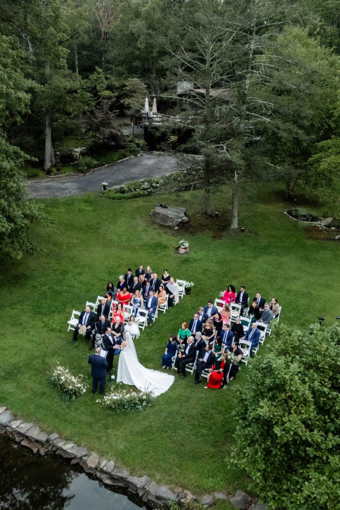 Aerial view of an outdoor wedding ceremony arranged beside a pond, with guests seated in a circular layout.