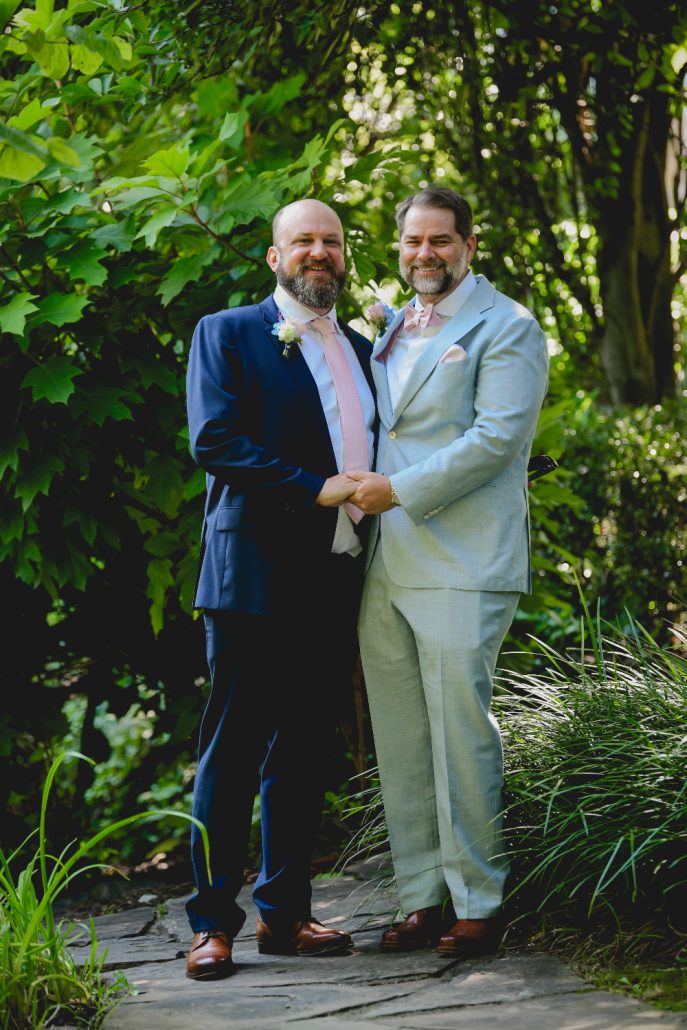 Grooms standing together and smiling in a lush garden setting, framed by greenery before their celebration.