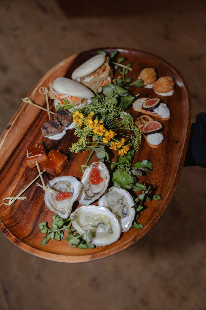 Wooden tray of assorted wedding hors d’oeuvres, including oysters, bao buns, figs, and seasonal garnishes for multi day wedding experiences.